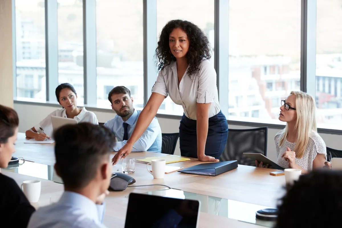 A woman stands at a conference table, embodying mindful leadership as she addresses her colleagues, blending traditional styles with a modern touch to enhance employee productivity in the office setting.