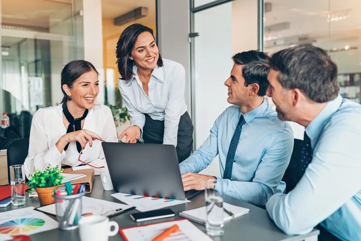 Four people in a meeting room are smiling and discussing around a laptop, with charts and a plant on the table, embracing mindful leadership to boost employee productivity.