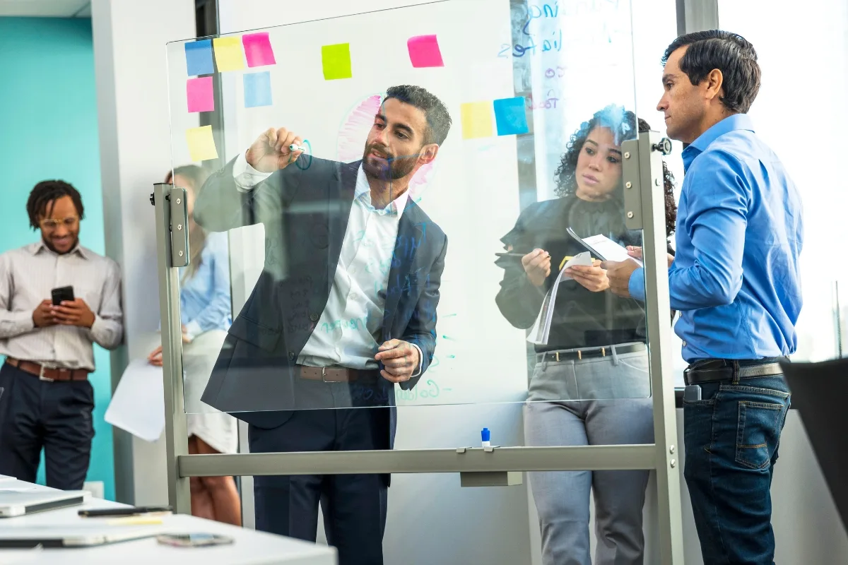 During a meeting focused on mindful leadership, a man writes on a transparent board, with three colleagues observing and taking notes to enhance employee productivity.