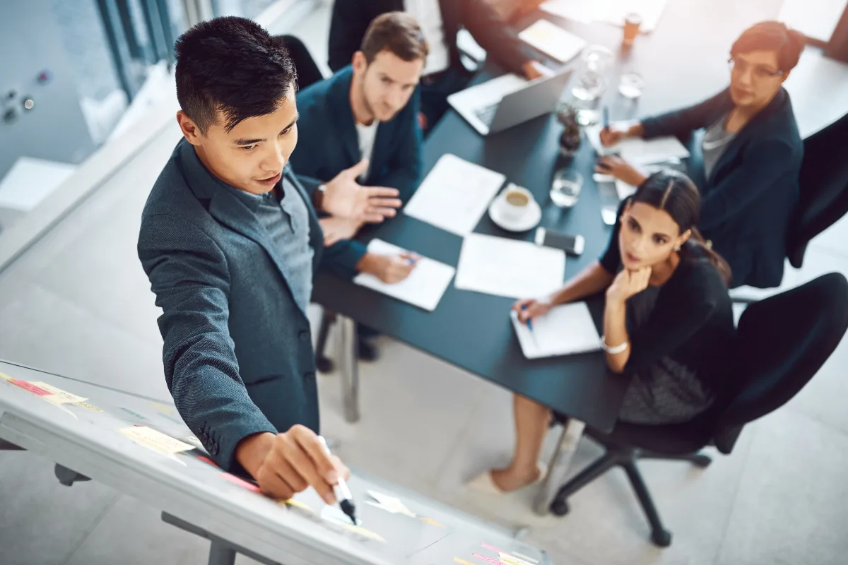 A man in a suit presents at a whiteboard, embodying mindful leadership as he engages a group of four people seated at a table with documents, laptops, and coffee cups. This approach fosters enhanced employee productivity during their collaborative session.