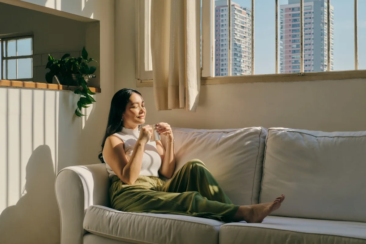 A woman sits on a white sofa in a sunlit room, embodying mindful productivity as she savors her mug of tea. She wears a white top and green pants, gazing at the tall buildings through the window. A potted plant graces the windowsill.