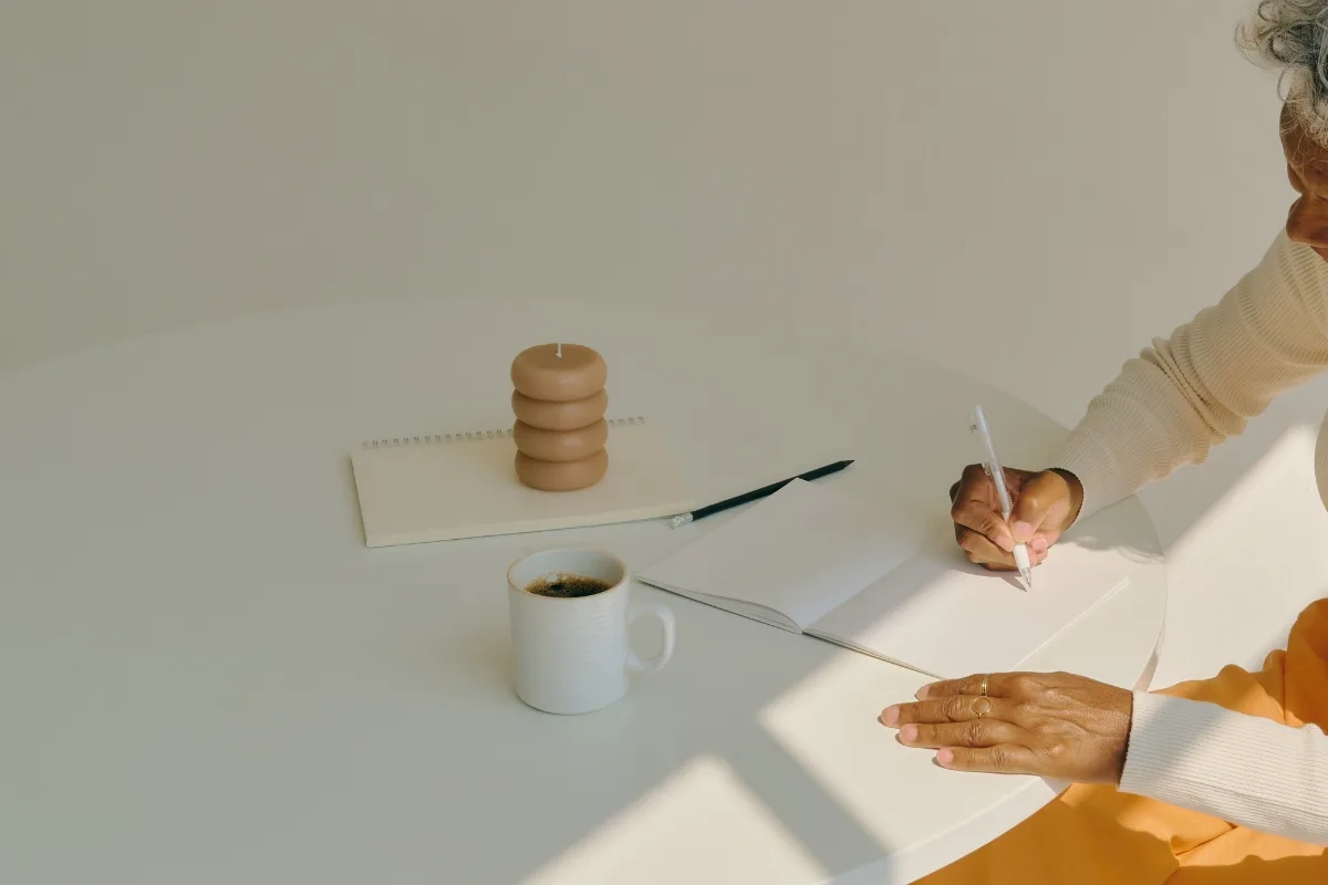 A person focuses on mindful productivity, writing in a notebook at a round table adorned with a steaming coffee mug, a stacked candle, and another notebook.