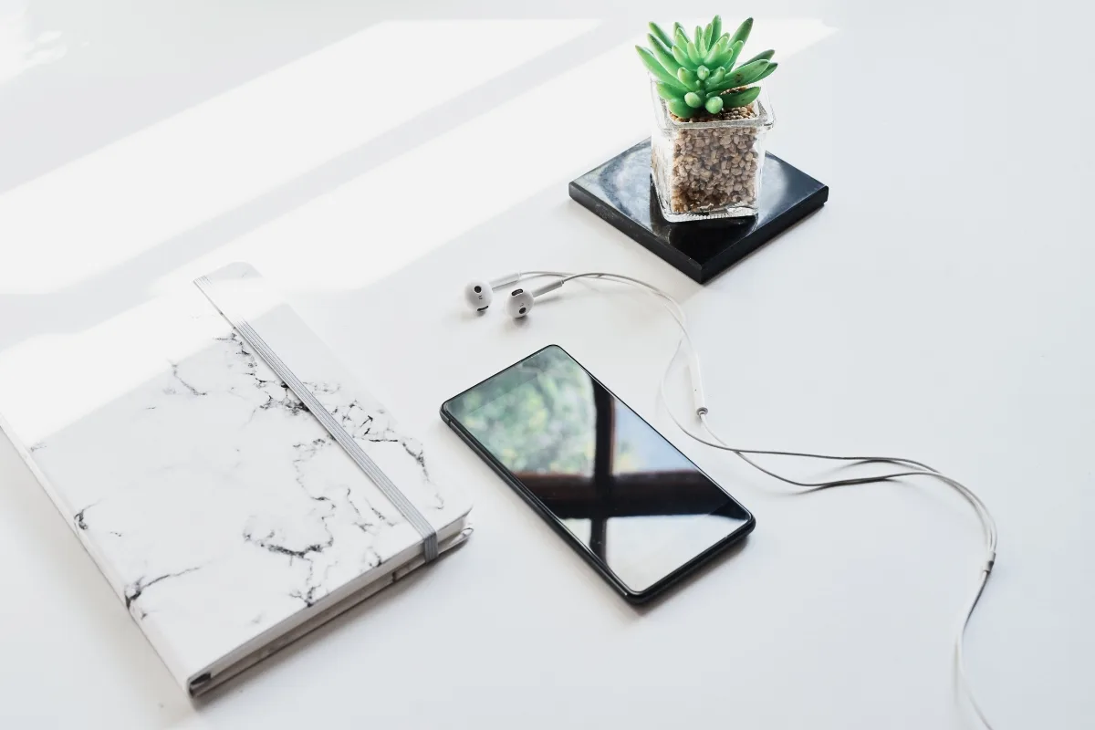 Smartphone with earphones, a closed marble-patterned notebook, and a small potted plant on a white surface create an atmosphere of mindful productivity.