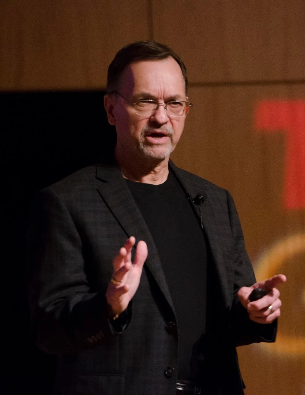 Dennis Buttimer, in a black shirt and blazer, speaks passionately on stage, gesturing with his hands.