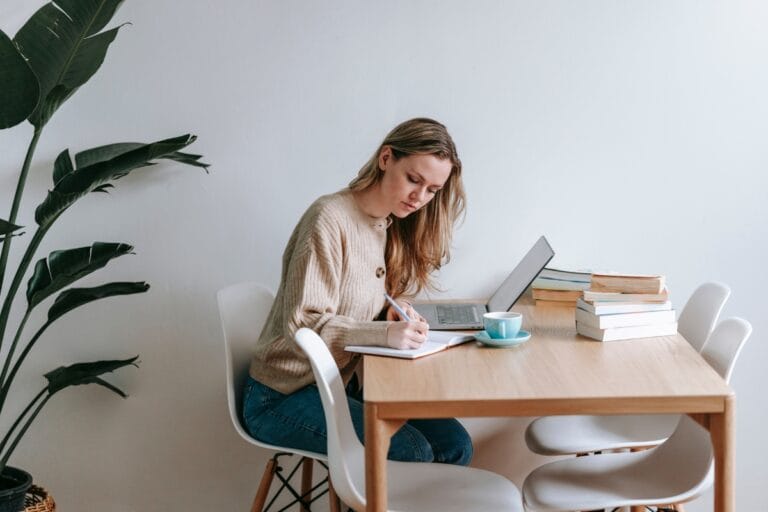 A woman sits at a table, staying focused at work with her laptop and notebook. A stack of books and a steaming cup keep her company, while a large plant adds a touch of nature to the corner.