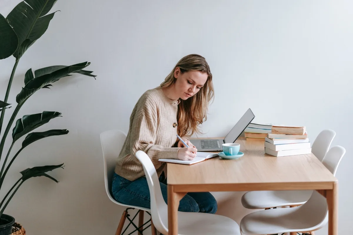 A woman sits at a table, staying focused at work with her laptop and notebook. A stack of books and a steaming cup keep her company, while a large plant adds a touch of nature to the corner.