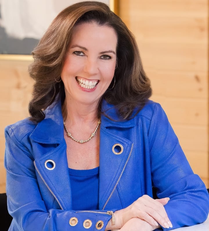 Woman with shoulder-length brown hair wearing a bright blue jacket and necklace, smiling and sitting at a table in a wood-paneled room, ready to discuss Counseling Services with warmth and professionalism.