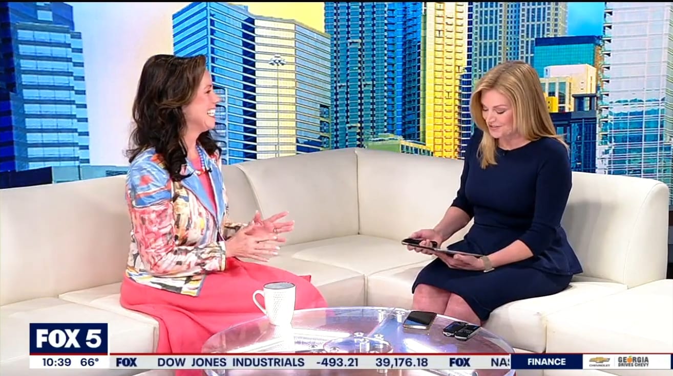 Two women sit on a white couch in a TV studio, one holding a tablet during a consulting segment. A FOX 5 news banner and cityscape backdrop are visible in the background.
