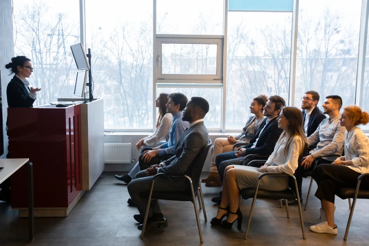 A group of people in business attire sit in rows, attentively listening to a speaker at a lectern. The bright room, with its large windows, provides the perfect setting for insights on executive coaching that might be right for you.