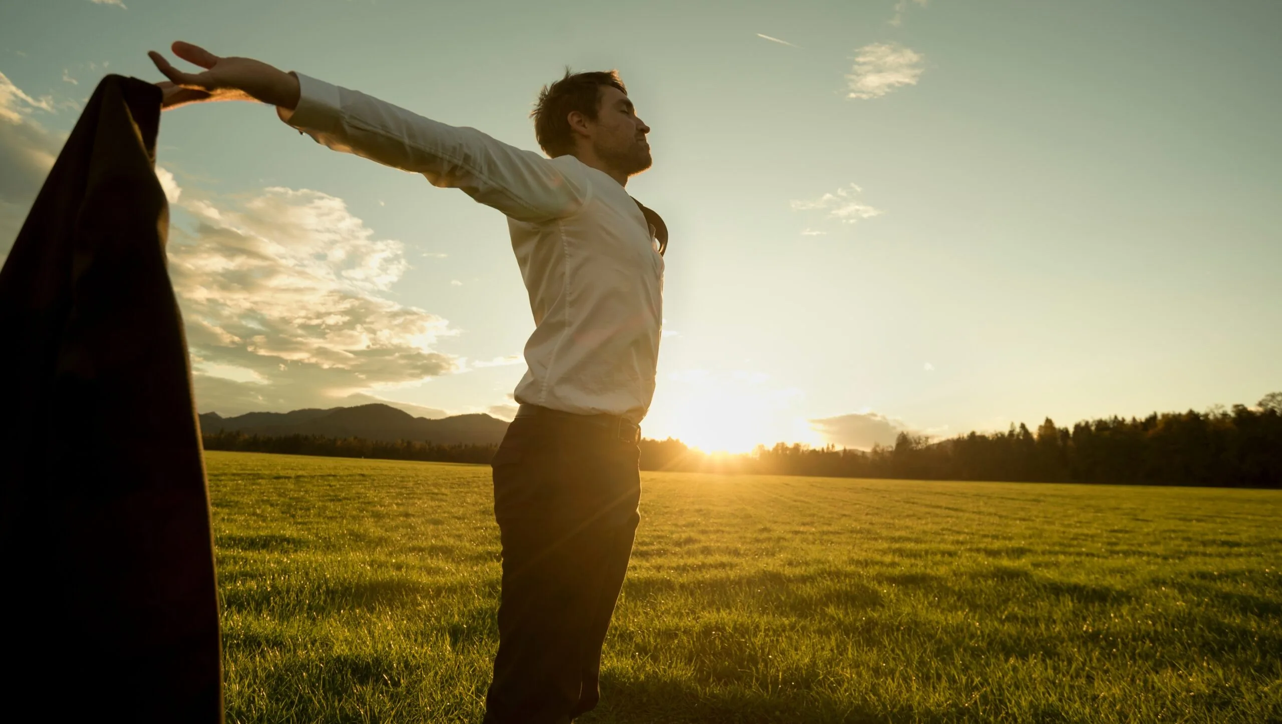 A person stands in a grassy field at sunset with arms outstretched, holding a jacket in one hand, embracing the sense of freedom and balance inspired by Mind-Body Psychotherapy.