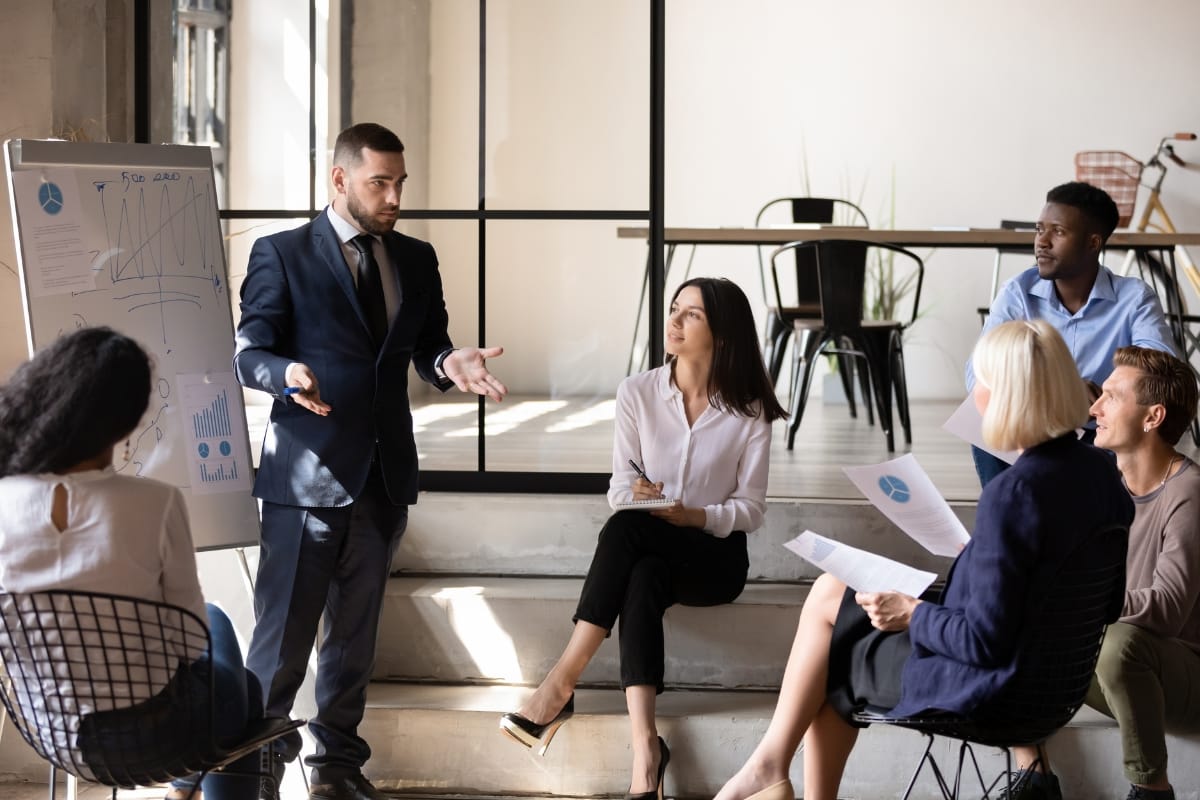 A man in a suit presents data on a flipchart to five seated colleagues in a modern office, illustrating the impact of Career Growth Coaching on team development.