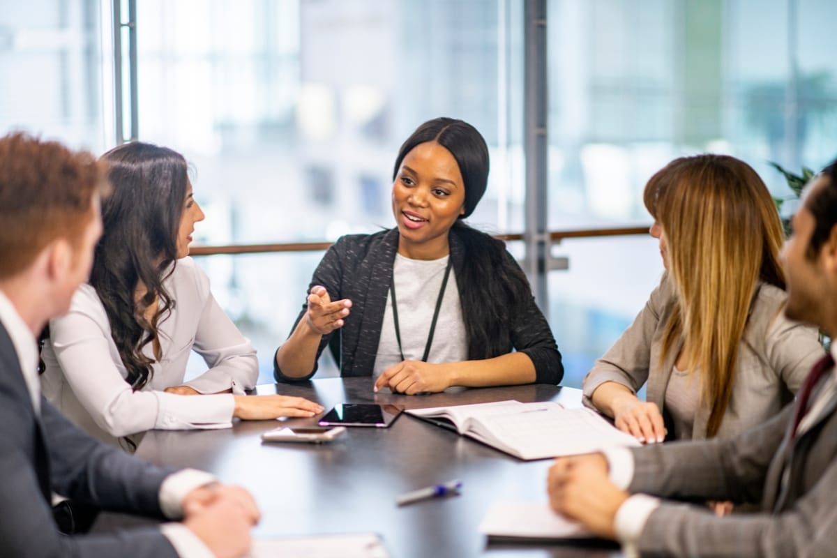Five professionals sit at a conference table having a discussion, with one woman in the center speaking and gesturing about Career Growth Coaching while others listen attentively.