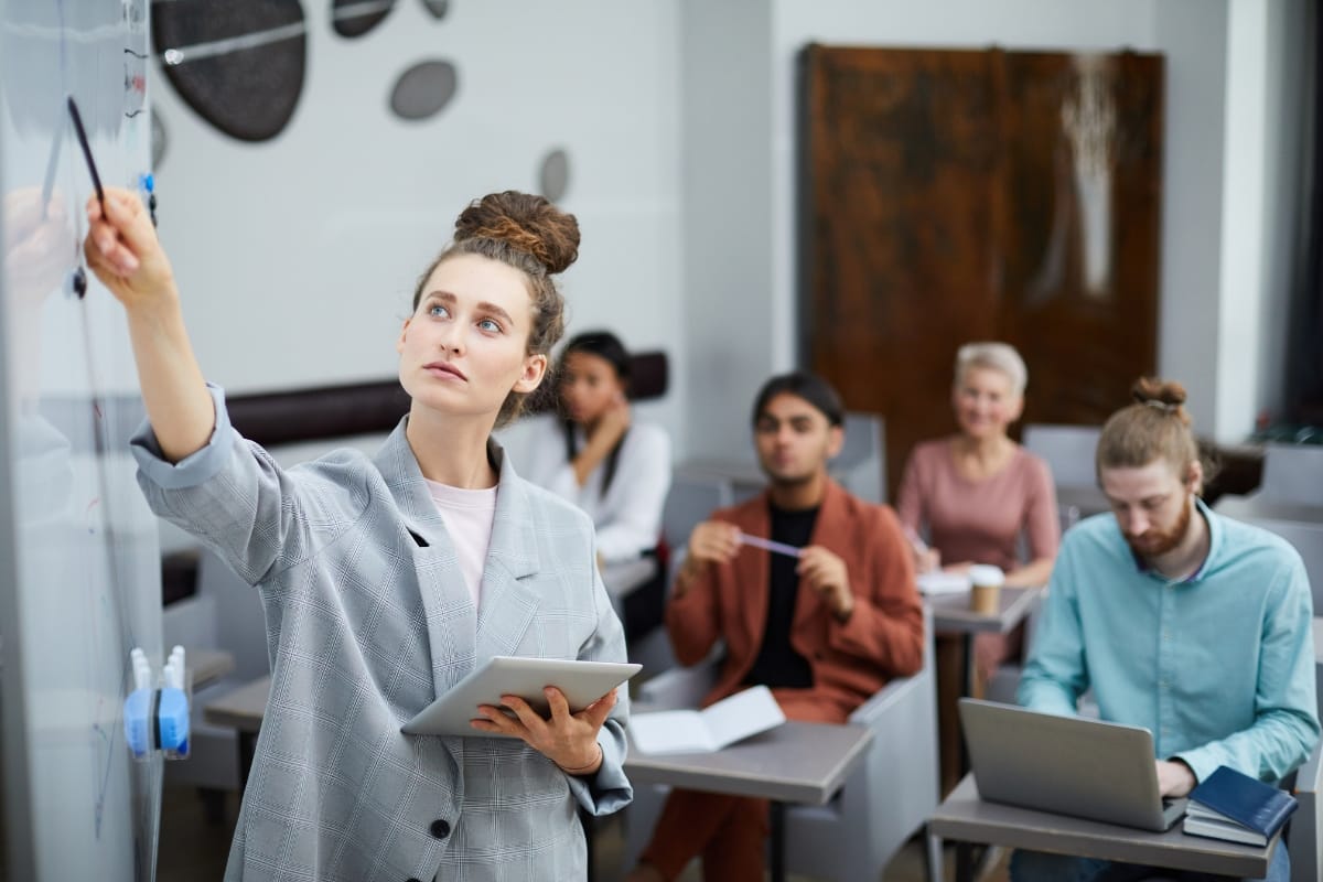 A woman leads a Career Growth Coaching session, writing on a whiteboard while holding a tablet, as four adults seated behind her take notes and use laptops in the classroom.