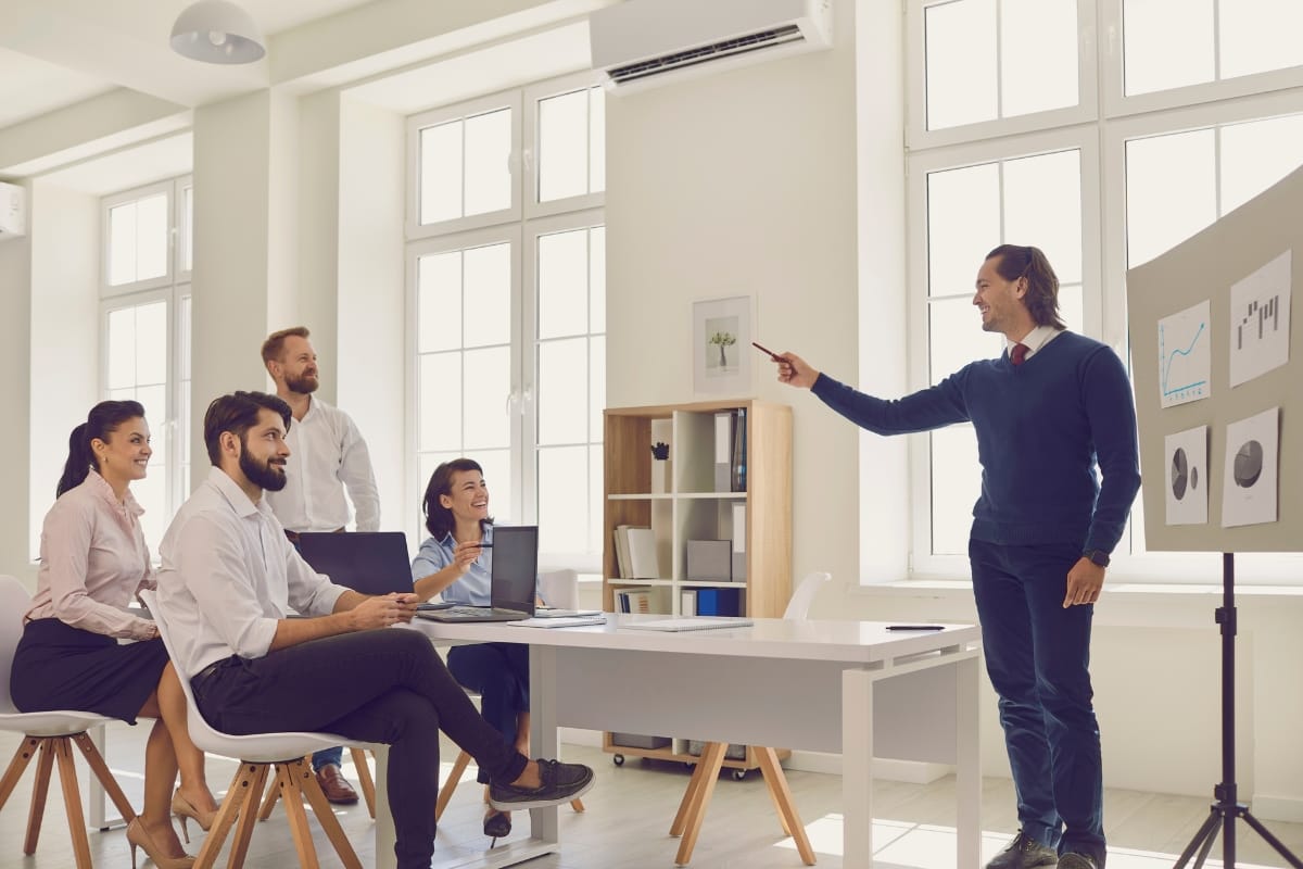 A man stands and gestures toward a presentation board with charts while four colleagues sit and watch, engaging in a Career Growth Coaching session in a bright office setting.