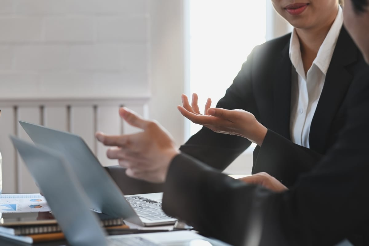 Two people in business attire sit at a table with laptops, engaged in a Career Growth Coaching discussion, using hand gestures to communicate.