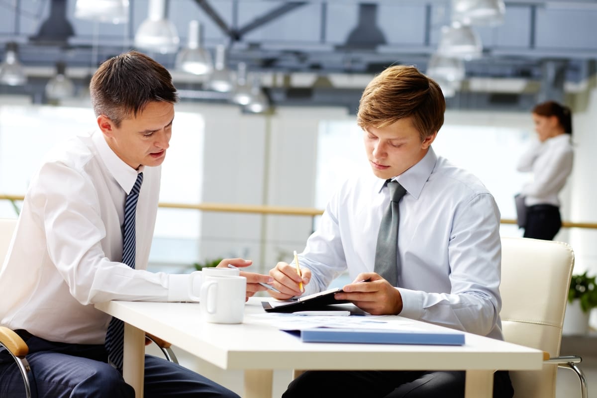 Two men in business attire sit at a table in an office, discussing documents and taking notes—perhaps during a Career Growth Coaching session—as a woman stands in the background.