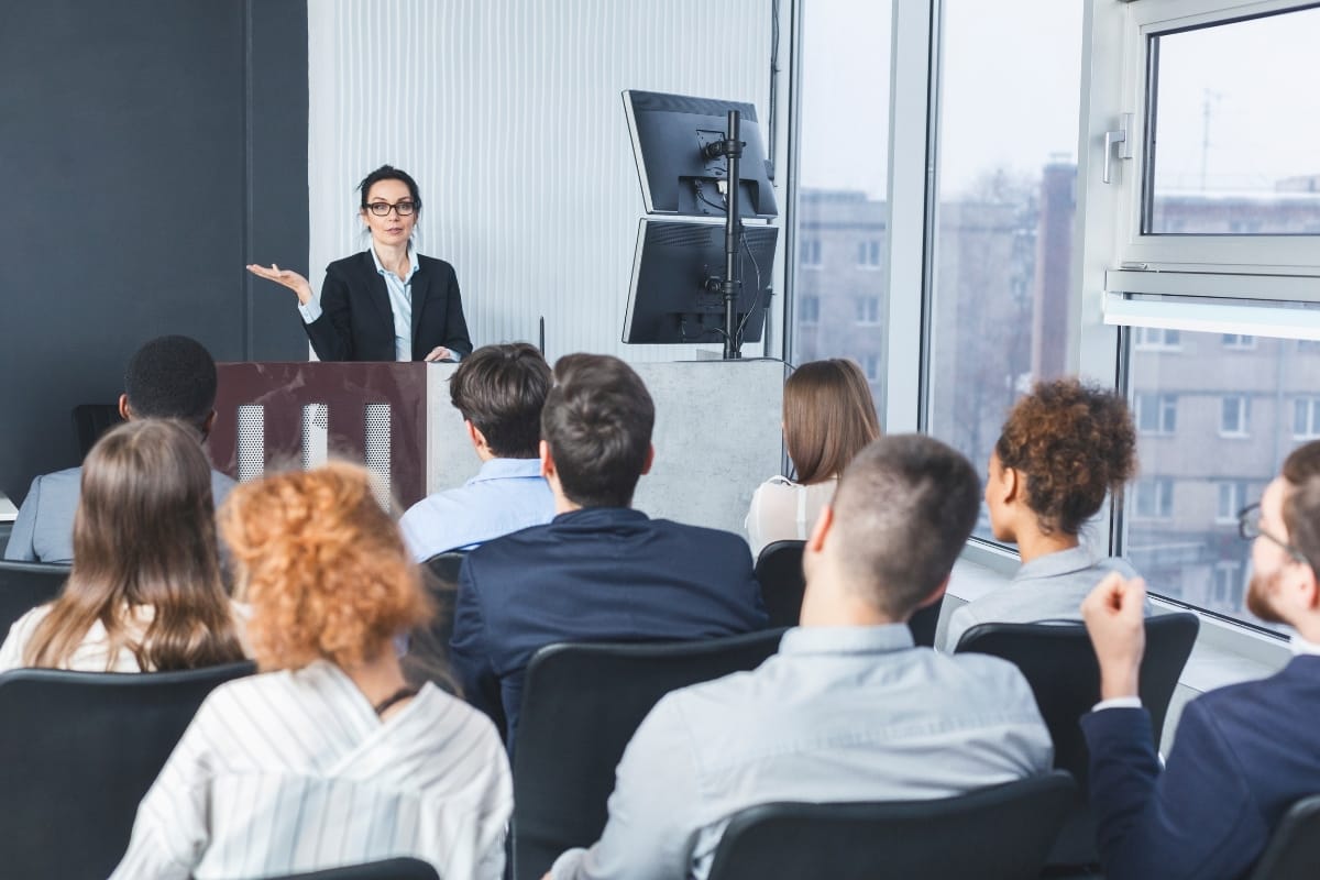 A woman stands at a podium giving a Career Growth Coaching presentation to a seated audience in a modern conference room with large windows and computer monitors.