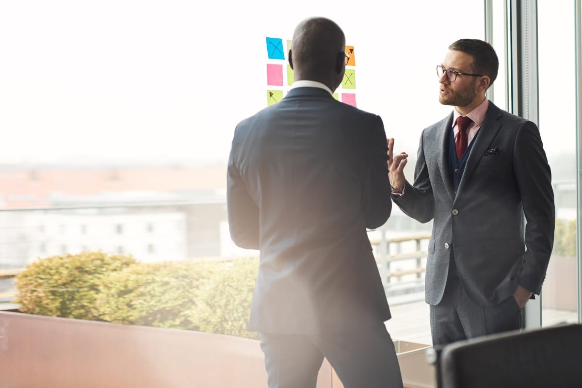 Two men in business suits stand by a window, discussing colorful sticky notes on glass—it’s a modern office setting focused on Career Growth Coaching.