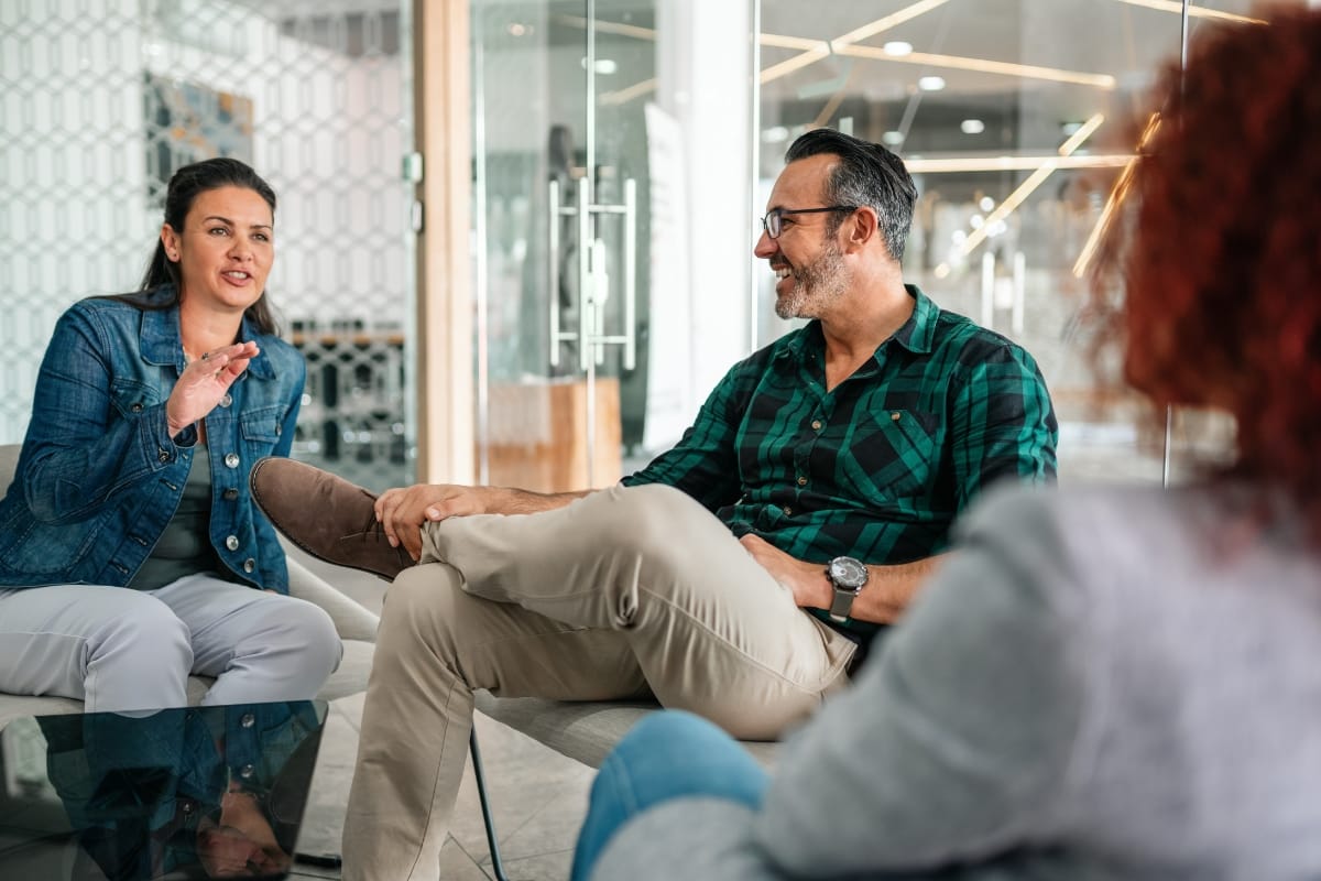 Three people sit in a modern office, having a conversation. The woman on the left gestures while speaking about Career Growth Coaching, and the man in the middle listens and smiles.