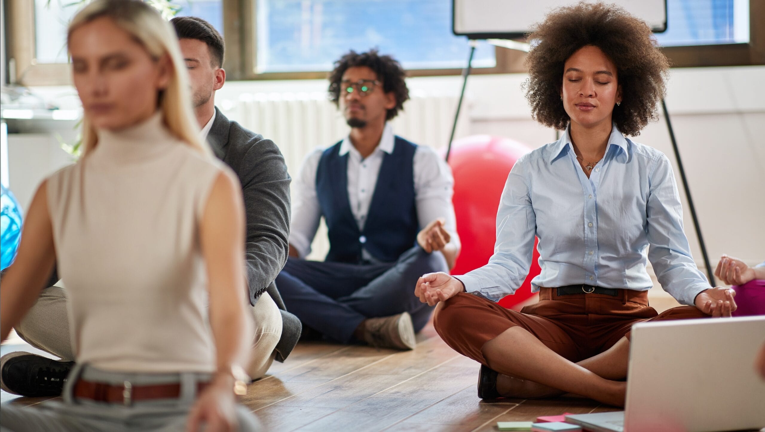A group of people sit cross-legged on the floor in an office, meditating with eyes closed and hands resting on their knees, practicing techniques inspired by Mind-Body Psychotherapy.