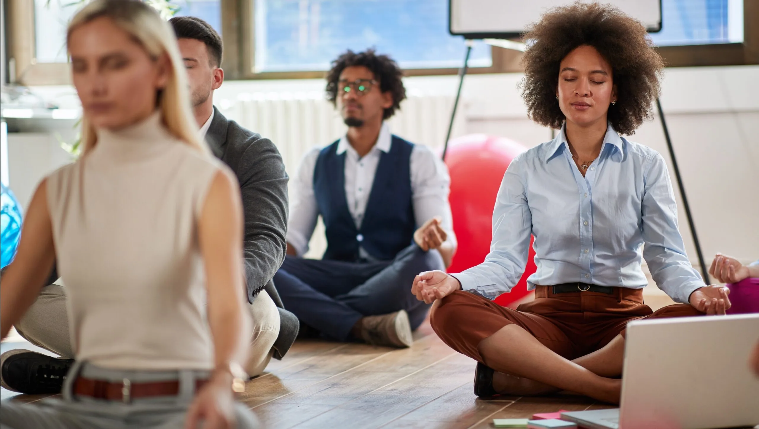 A group of people sit cross-legged on the floor in an office, meditating with eyes closed and hands resting on their knees, practicing techniques inspired by Mind-Body Psychotherapy.