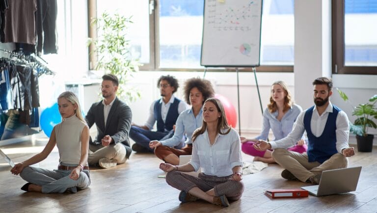 A group of people in business attire sit cross-legged on the floor meditating in an office, with a flip chart and laptop visible, embracing Mind-Body Psychotherapy techniques for workplace well-being.