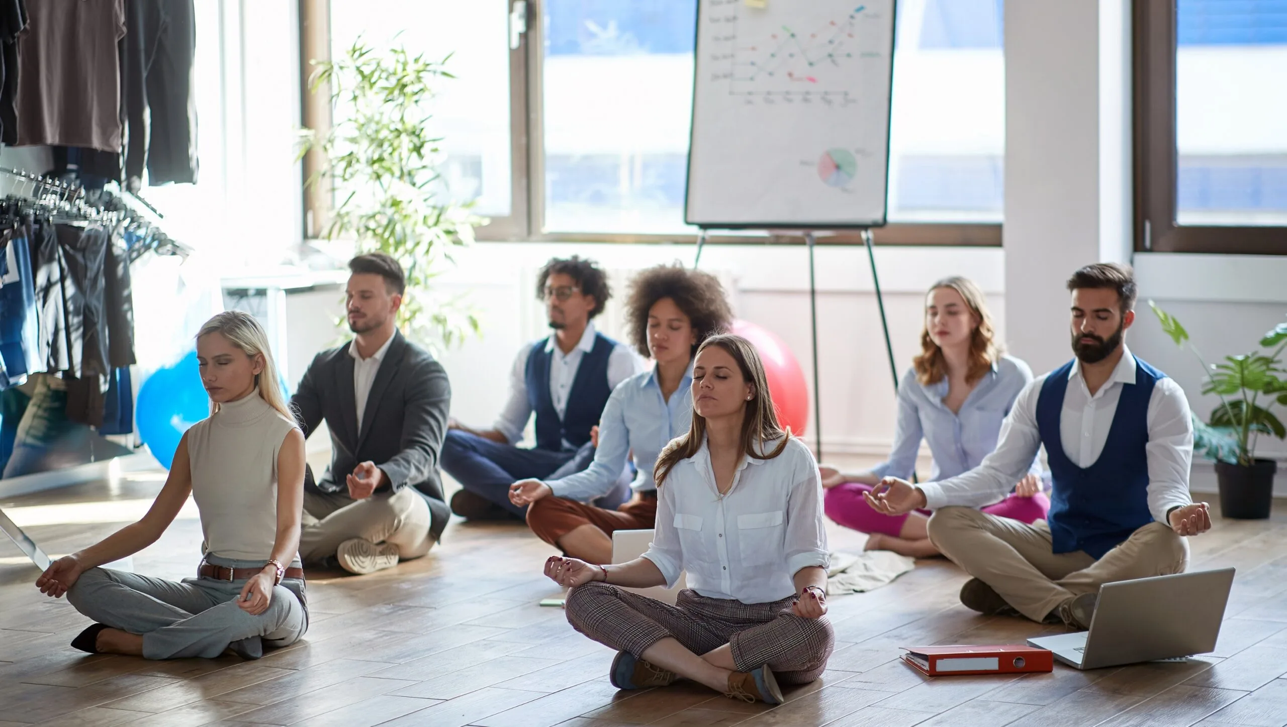 A group of people in business attire sit cross-legged on the floor meditating in an office, with a flip chart and laptop visible, embracing Mind-Body Psychotherapy techniques for workplace well-being.