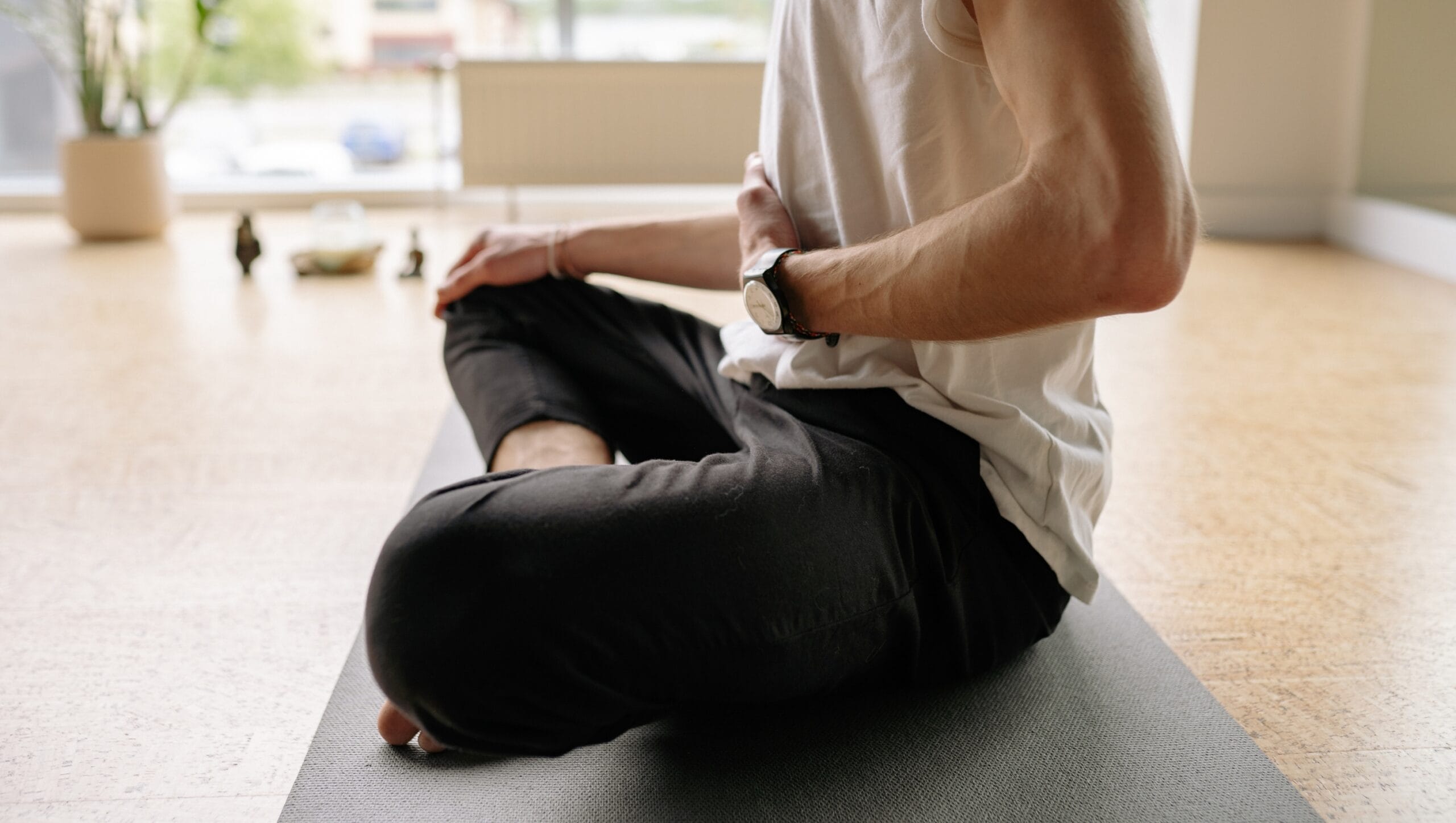 Person sitting cross-legged on a yoga mat in a bright room, wearing a watch and holding one hand to their abdomen, practicing mindful breathing—a calming moment inspired by Mind-Body Psychotherapy, with blurred objects in the background.