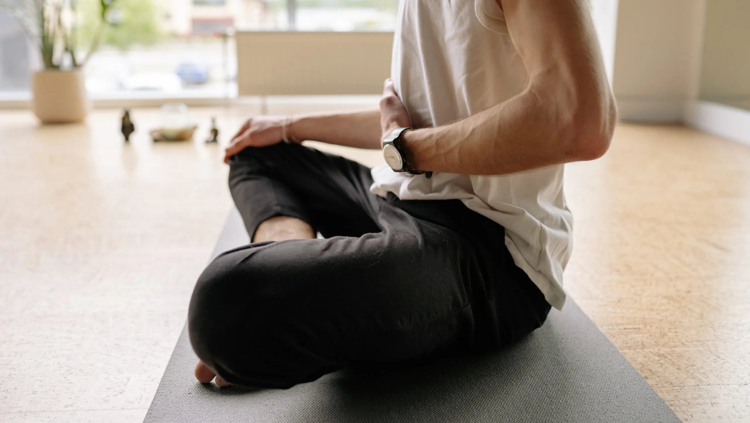 Person sitting cross-legged on a yoga mat in a bright room, wearing a watch and holding one hand to their abdomen, practicing mindful breathing—a calming moment inspired by Mind-Body Psychotherapy, with blurred objects in the background.