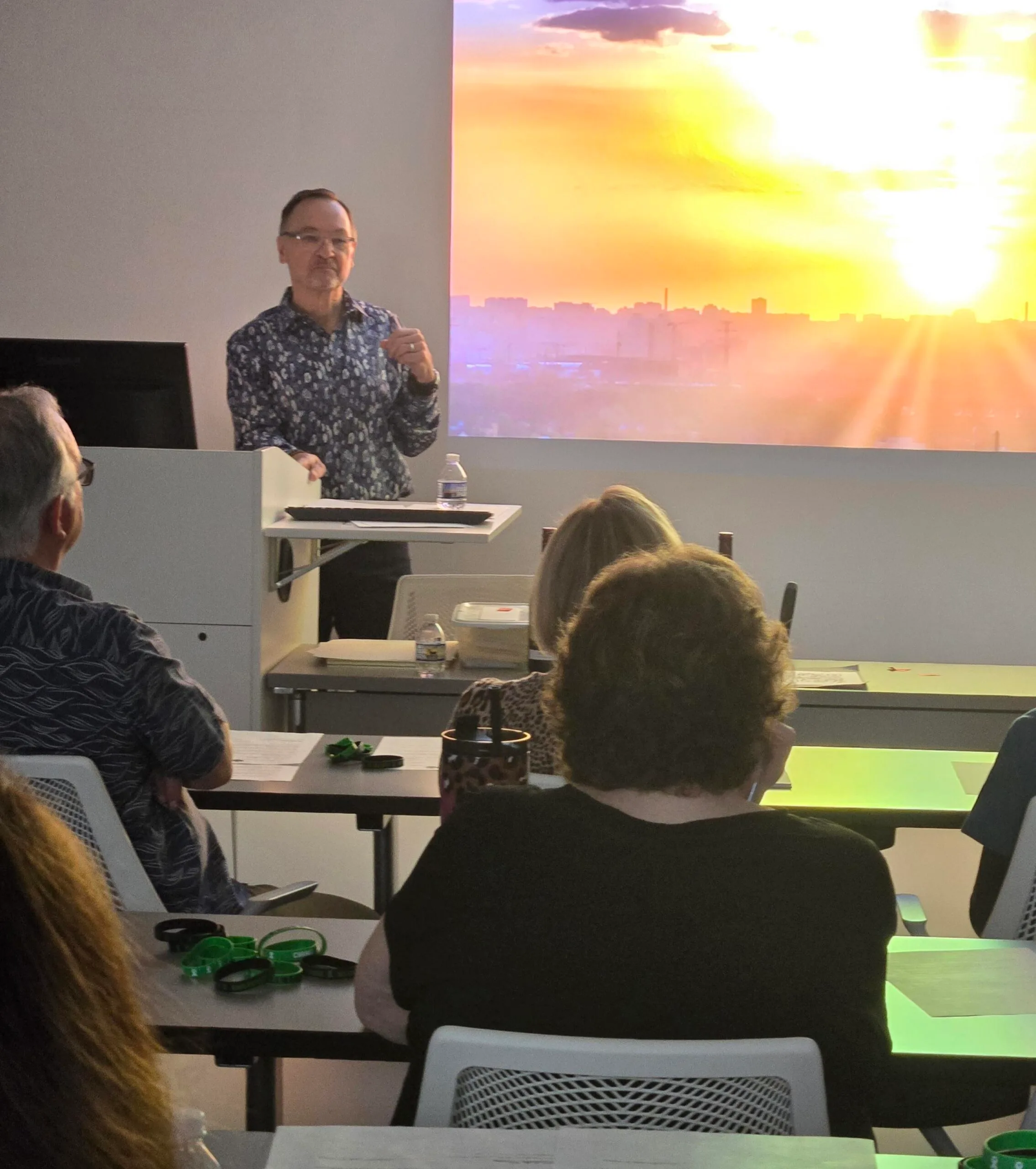 Dennis Buttimer stands at a podium giving a presentation to a seated audience, with a large projected image of a city skyline at sunset displayed behind him.