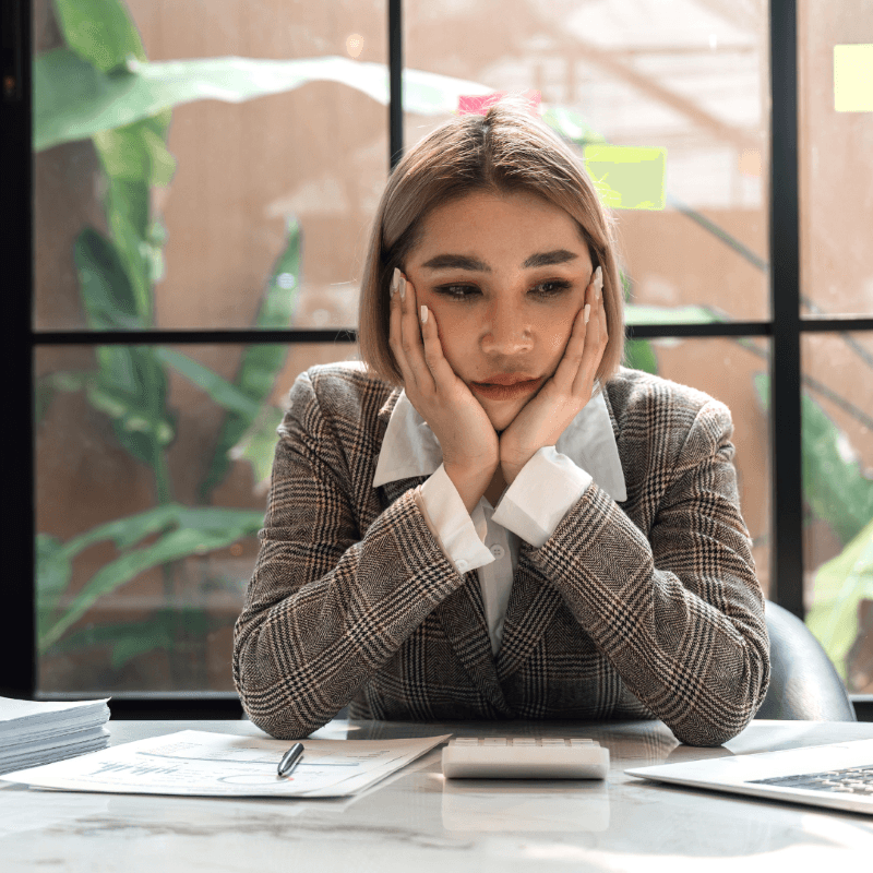 A woman in a plaid blazer sits at a desk with papers and a calculator, resting her face in her hands and looking thoughtful, perhaps reflecting on employee motivation and business growth.