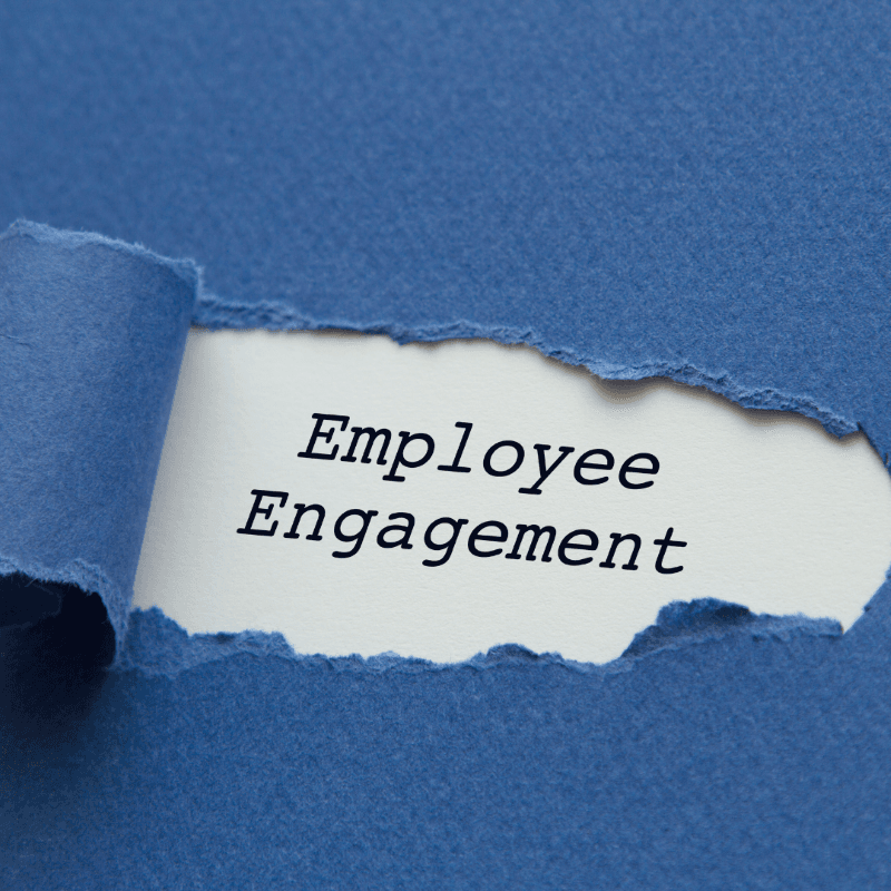 A piece of blue paper is torn away to reveal the words "Employee Engagement" typed on white paper underneath, symbolizing uncovering effective employee engagement strategies within a business.