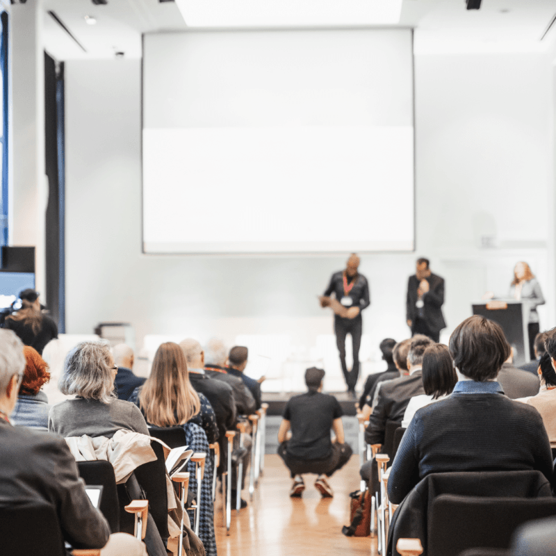 A group of people seated in an auditorium face a stage where three speakers stand in front of a projector screen, presenting insights on employee engagement.
