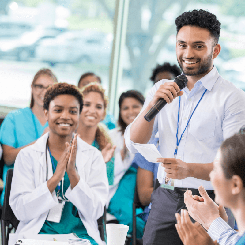 A man holding a microphone speaks to an audience of people, including medical professionals in white coats and scrubs, who are clapping and listening attentively, highlighting the importance of employee engagement.