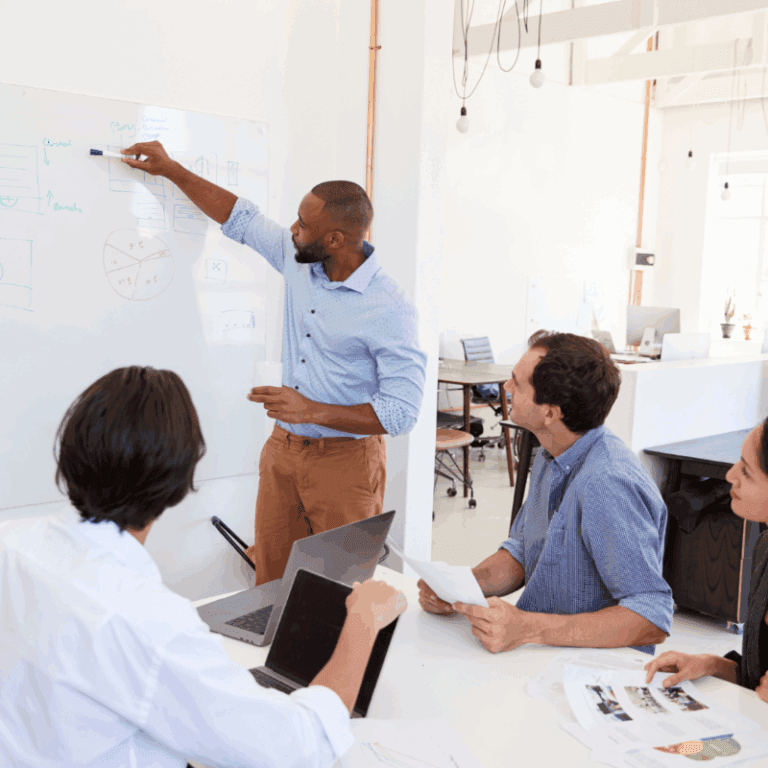 Man stands at a whiteboard explaining diagrams to three seated colleagues in a modern office setting, bolstering employee engagement as they review papers and laptops at the table.
