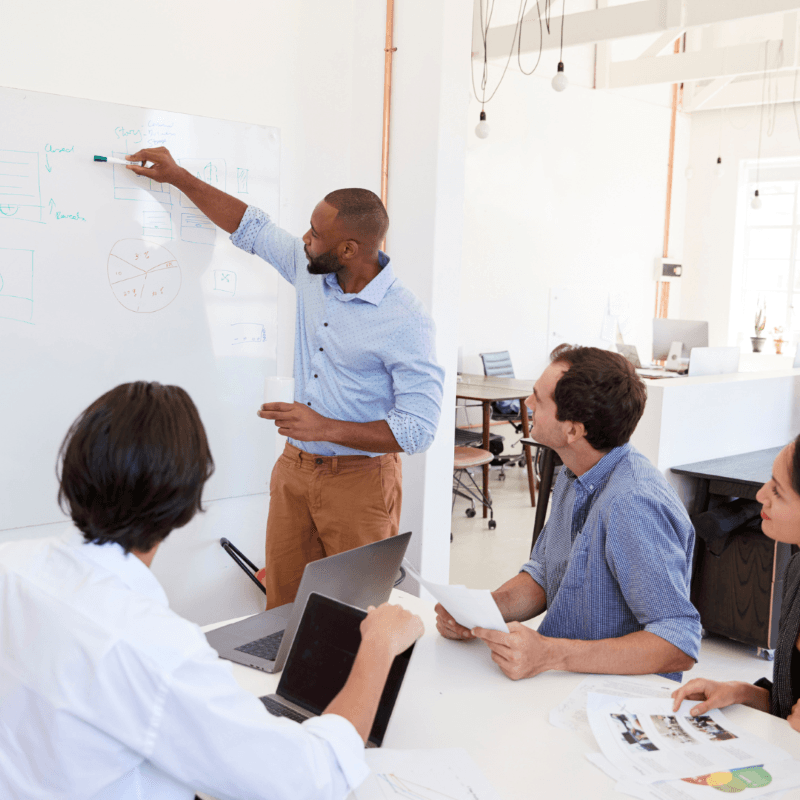 Man stands at a whiteboard explaining diagrams to three seated colleagues in a modern office setting, bolstering employee engagement as they review papers and laptops at the table.