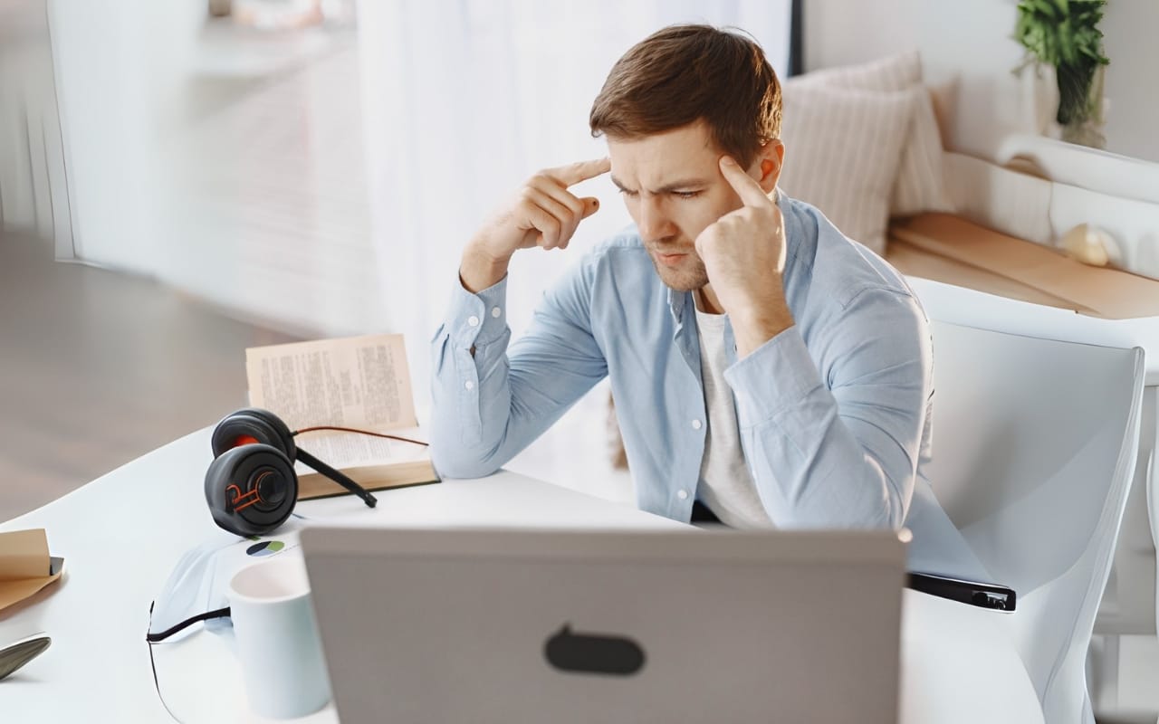 A man sits at a desk in front of a laptop, touching his temples and appearing focused or stressed. Surrounded by a headset, notebook, and book, he seeks productivity while navigating challenges—perhaps using effective ADHD tips.
