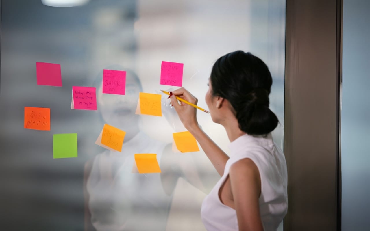 A woman writes on a sticky note attached to a glass wall filled with colorful notes, showcasing her productivity and organized approach.