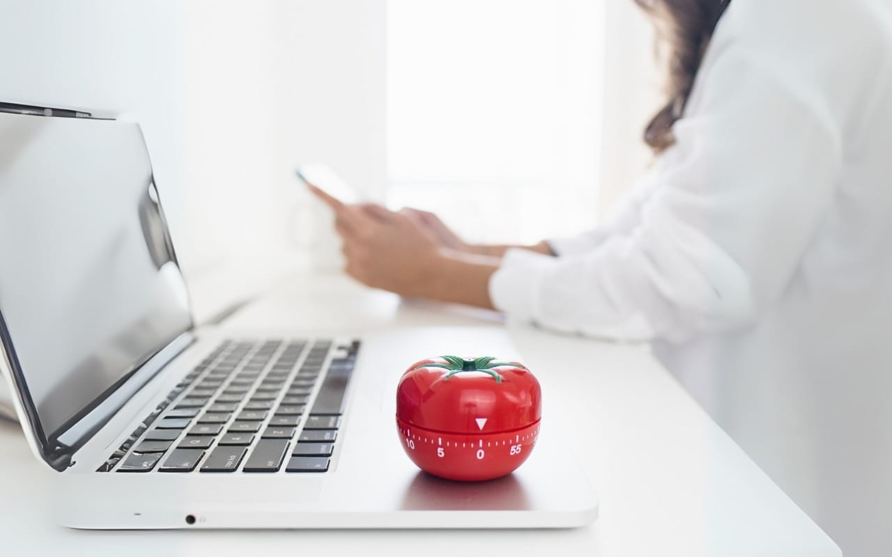 A red tomato-shaped kitchen timer sits on a laptop beside a person using a smartphone at a white desk, illustrating productivity and practical ADHD tips for effective time management.