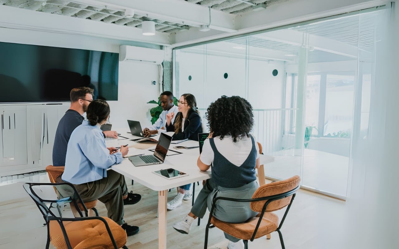 Five people sit at a conference table in a modern office, engaged in a productive meeting focused on leadership strategies, with laptops, documents, and notebooks on the table.