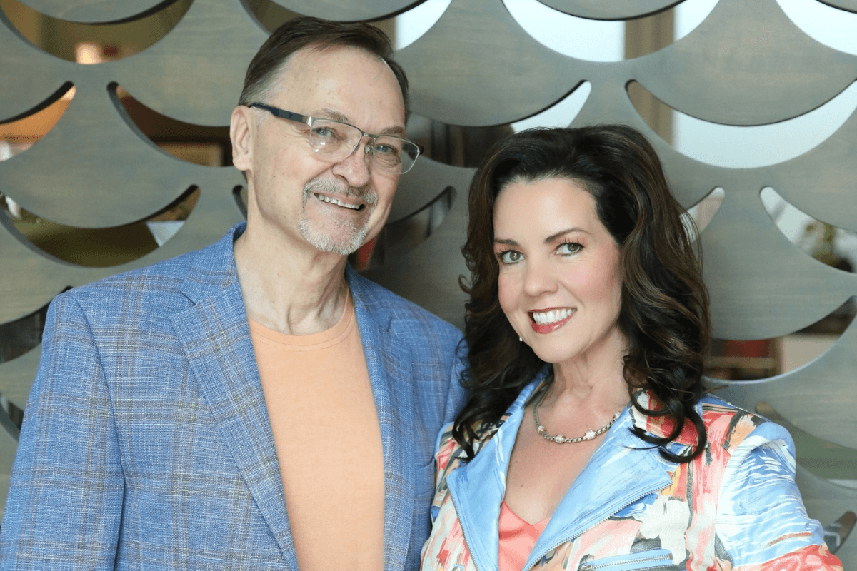A man in a blue plaid jacket and a woman in a floral jacket, both from the consulting team, stand side by side, smiling at the camera in front of a decorative wall.
