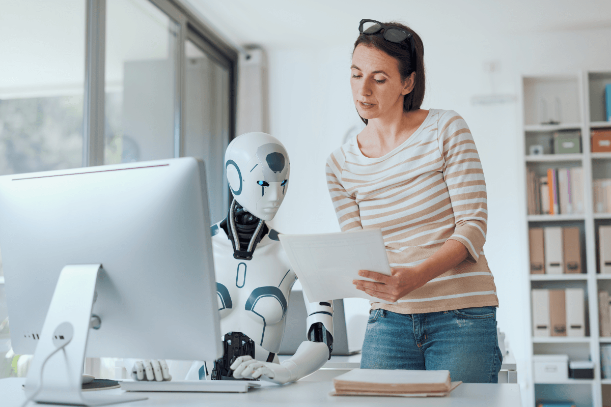 A woman stands next to a humanoid robot at a desk, showing it a document, as the robot tackles AI challenges while sitting in front of a computer monitor in a modern office setting.