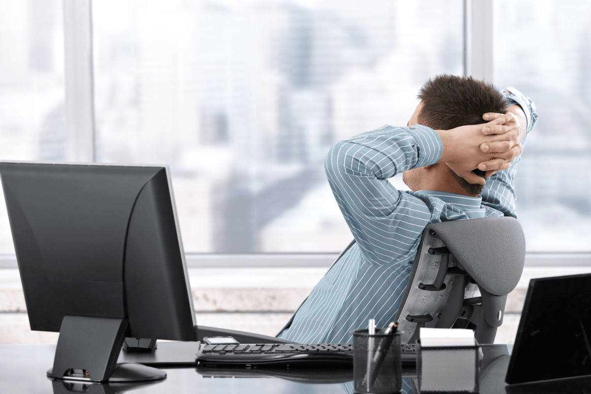 A person sits at a desk in an office, leaning back in their chair with hands clasped behind their head, embodying presence and mindful leadership as they gaze out large windows.