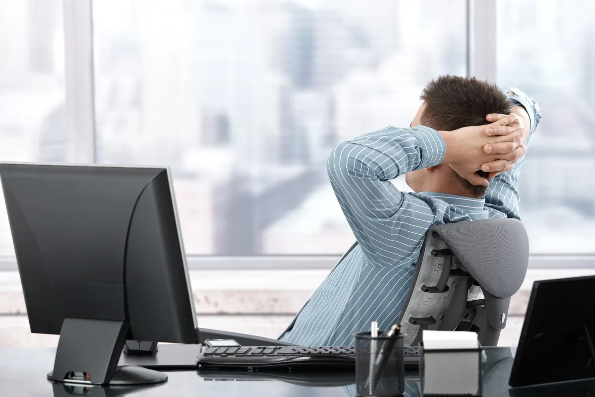 A person sits at a desk in an office, leaning back in their chair with hands clasped behind their head, embodying presence and mindful leadership as they gaze out large windows.