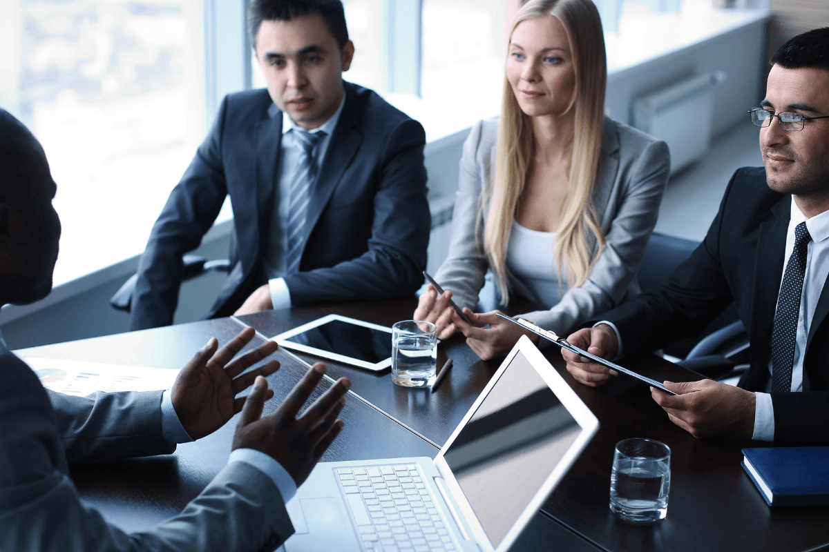 Four business professionals in formal attire sit around a conference table with laptops and documents, demonstrating mindful leadership and presence as they engage in a focused discussion.