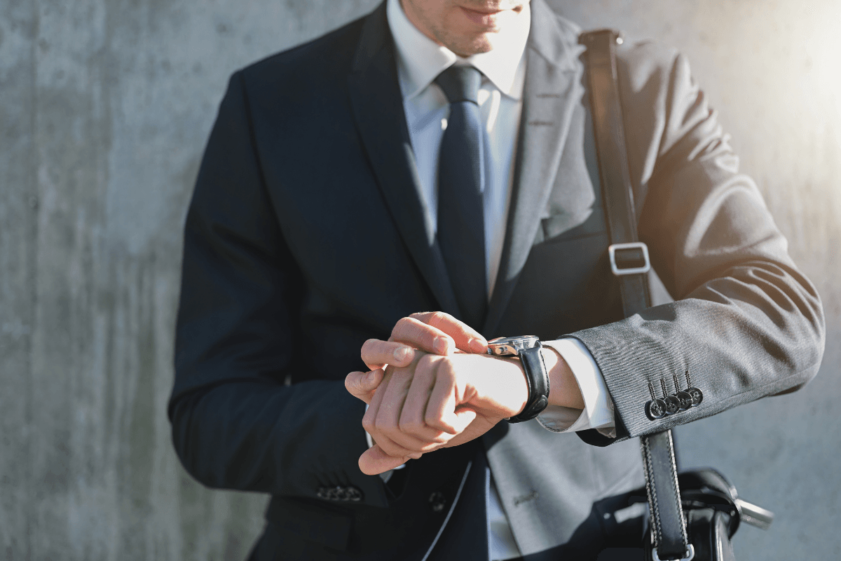 A man in a suit and tie checks the time on his wristwatch with presence and purpose, holding a bag over his shoulder while standing in front of a concrete wall.