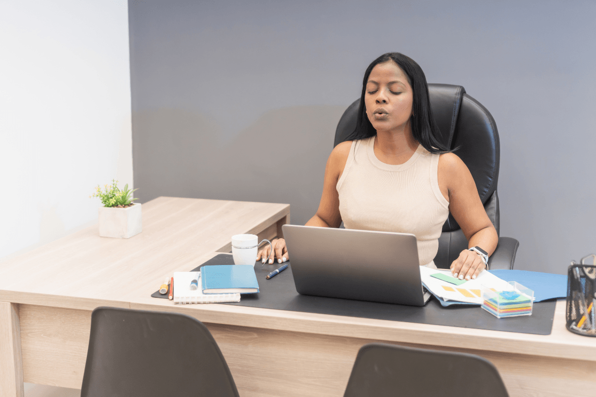 A woman sits at a desk with a laptop and documents, eyes closed and lips pursed as if practicing Presence and deep breathing—embracing Mindful Leadership to boost focus and balance during her workday.