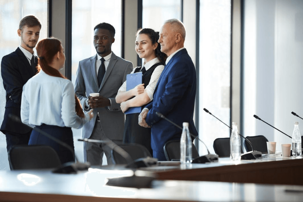 Five people in business attire stand and talk together in a conference room with large windows and microphones on the table, discussing practical skills that help them thrive at work.