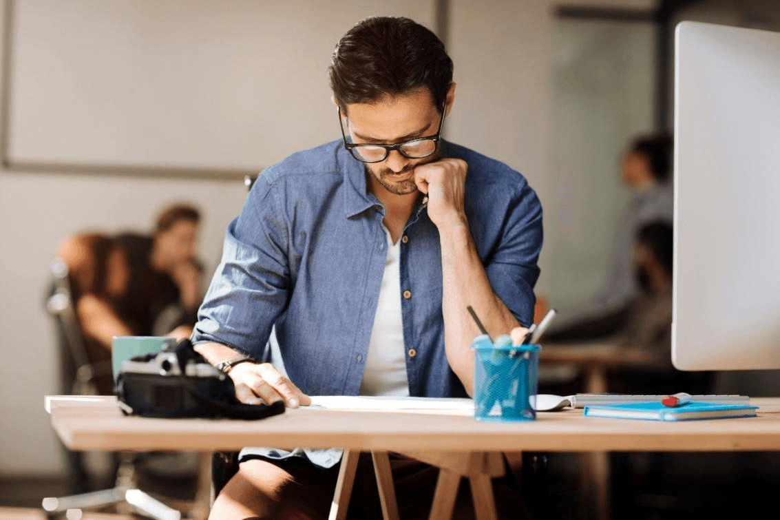 A man wearing glasses and a denim shirt sits at a desk in a modern office, looking down at papers with his hand on his chin—focused on developing practical skills to thrive at work.