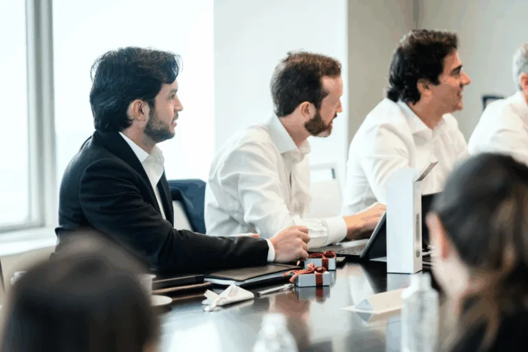 Three men in business attire sit at a conference table with laptops and papers, actively participating in a meeting.